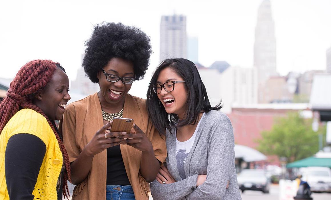 Three students presenting as women of color stand together smiling at phone with Kansas City skyline in background